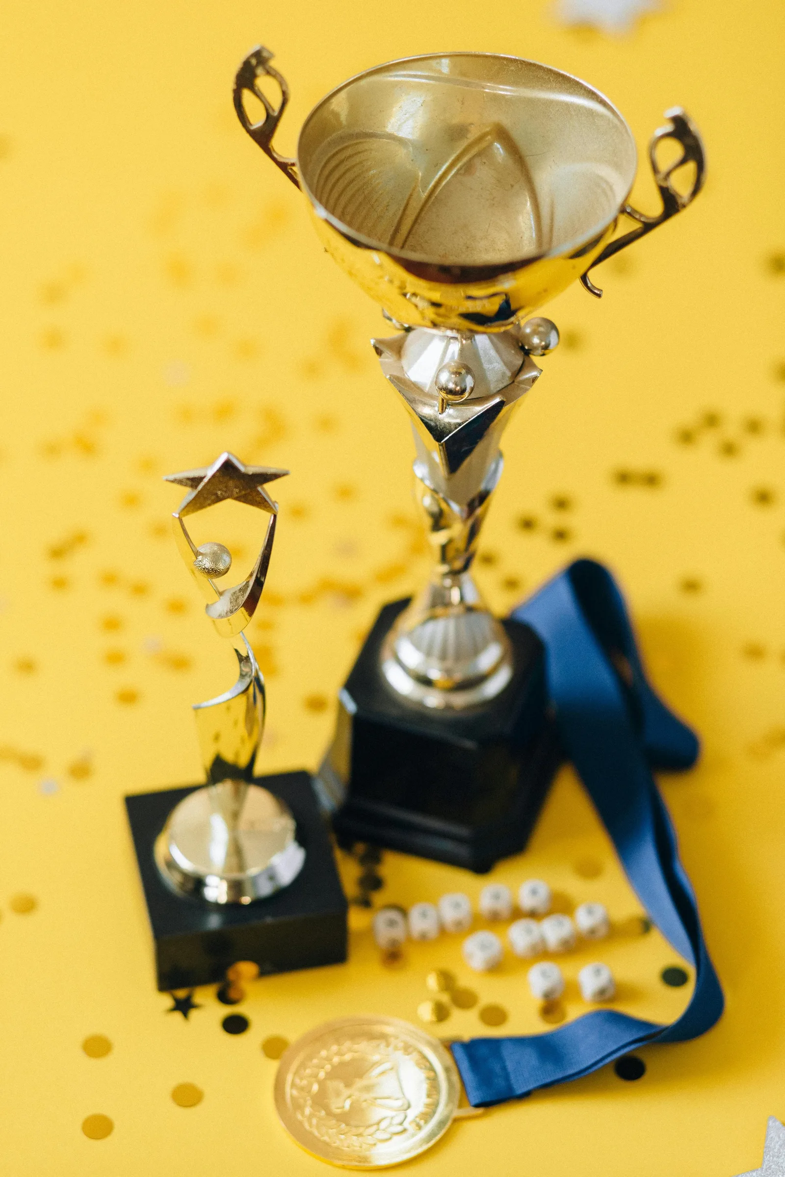 A trophy, a small star-shaped award, and a medal with ribbon sit among gold confetti on a yellow background.