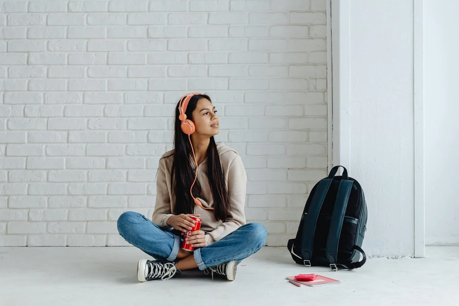 A teenage girl wearing pink headphones sits cross-legged on the floor against a white brick wall, next to a backpack.
