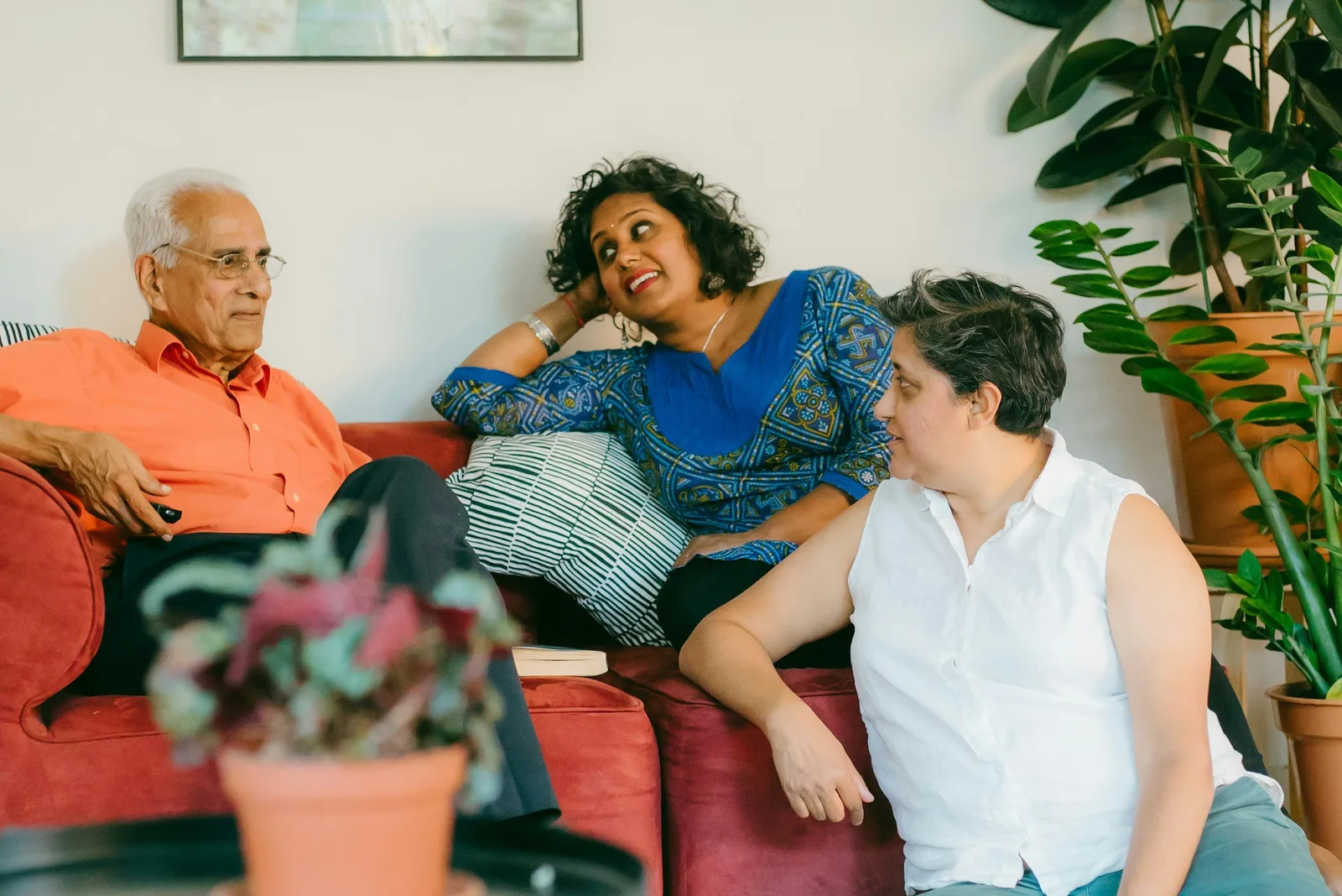 An older man, a woman, and a younger person sit together on a couch talking, with houseplants nearby.