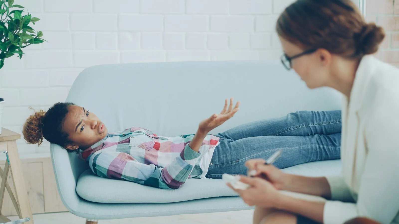 A girl reclines on a couch gesturing while talking with a therapist who takes notes.