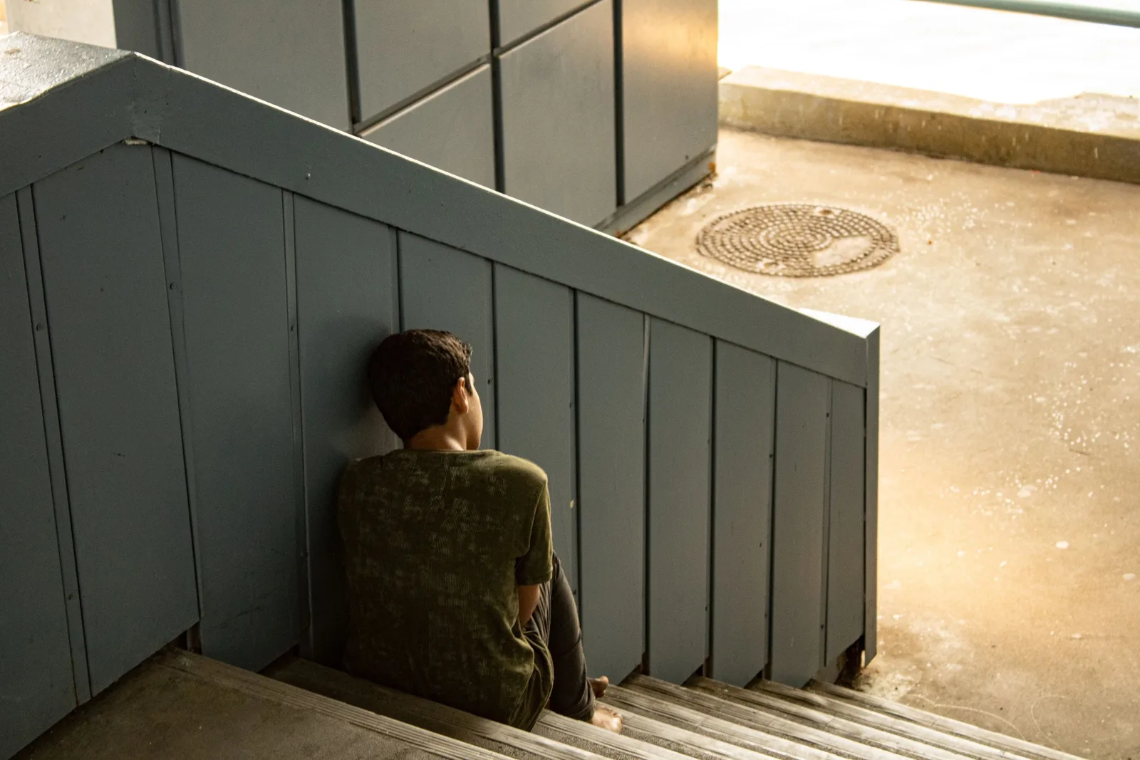 A boy sits alone on outdoor steps with his back to the camera, knees pulled up.