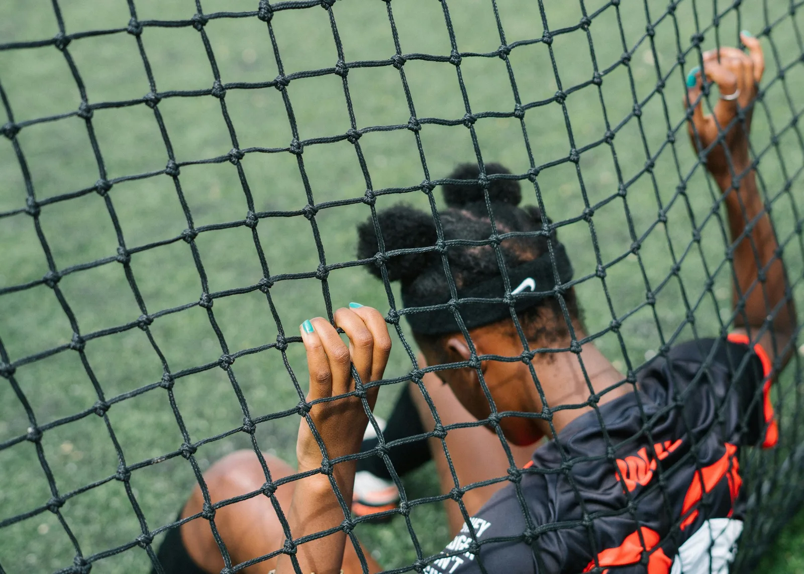 A young athlete peers through a practice net on a sports field, resting against the mesh.