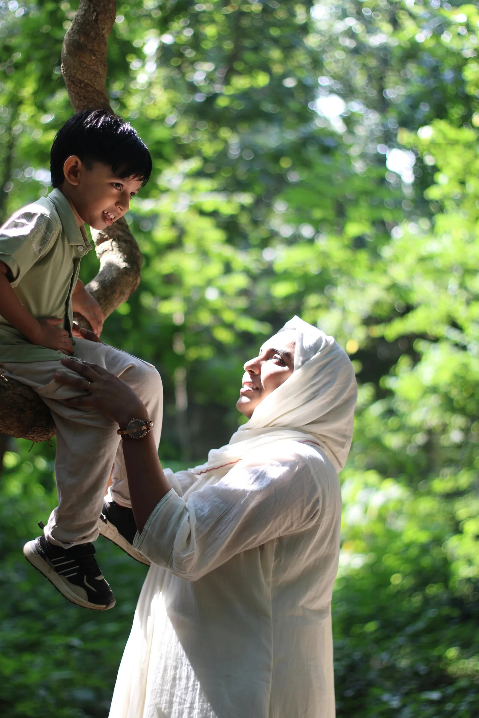 A woman in a white hijab lifts a laughing young boy up onto a tree branch in a green forest.