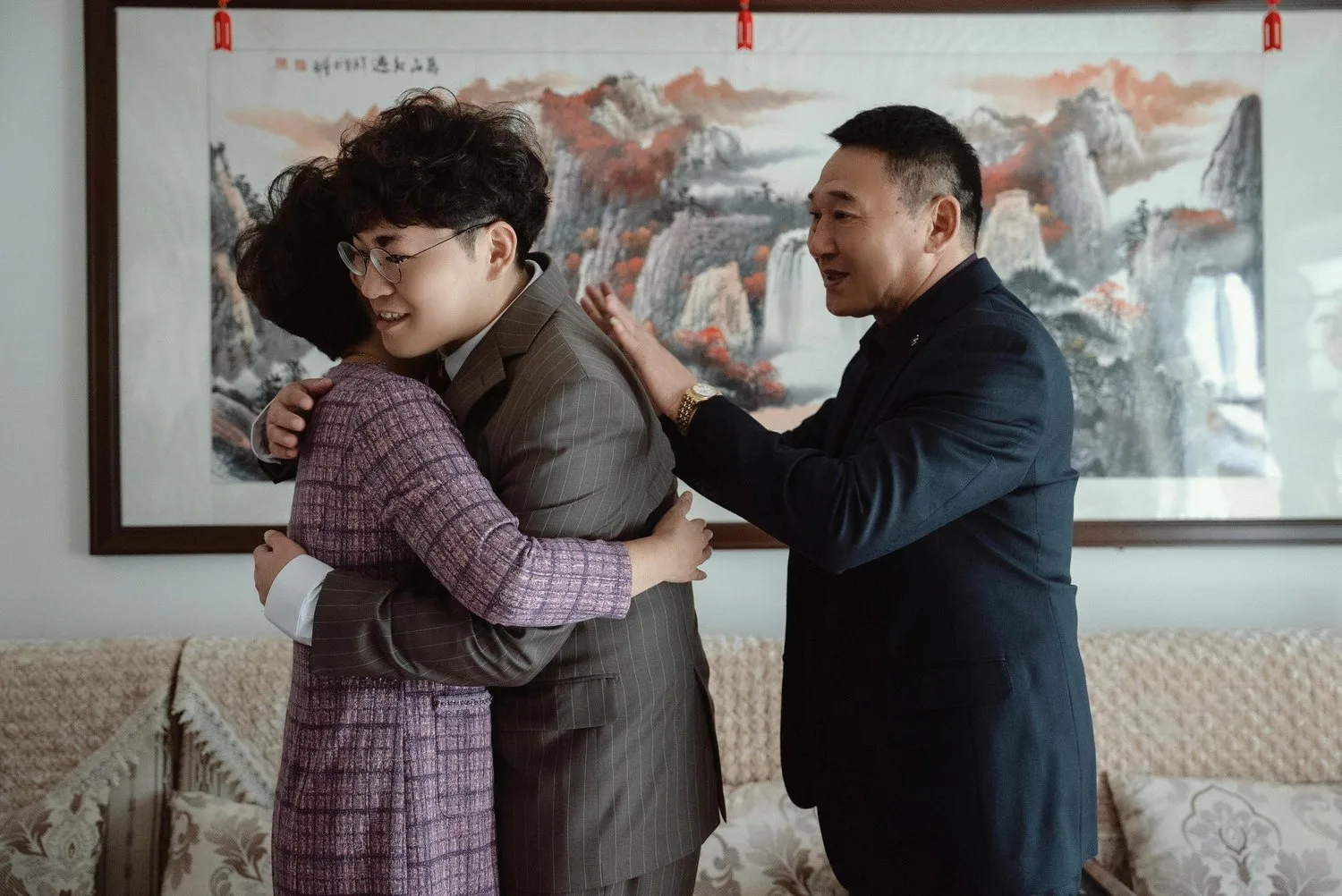 A woman is embraced by a man while an older man places his hands on the man’s shoulders, in front of a Chinese landscape painting.
