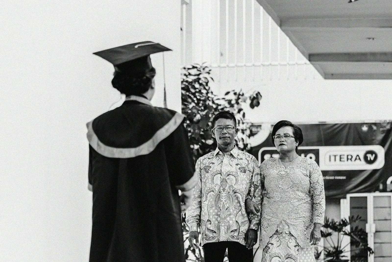 A graduate in cap and gown faces two smiling parents at a graduation ceremony, shown in black and white.
