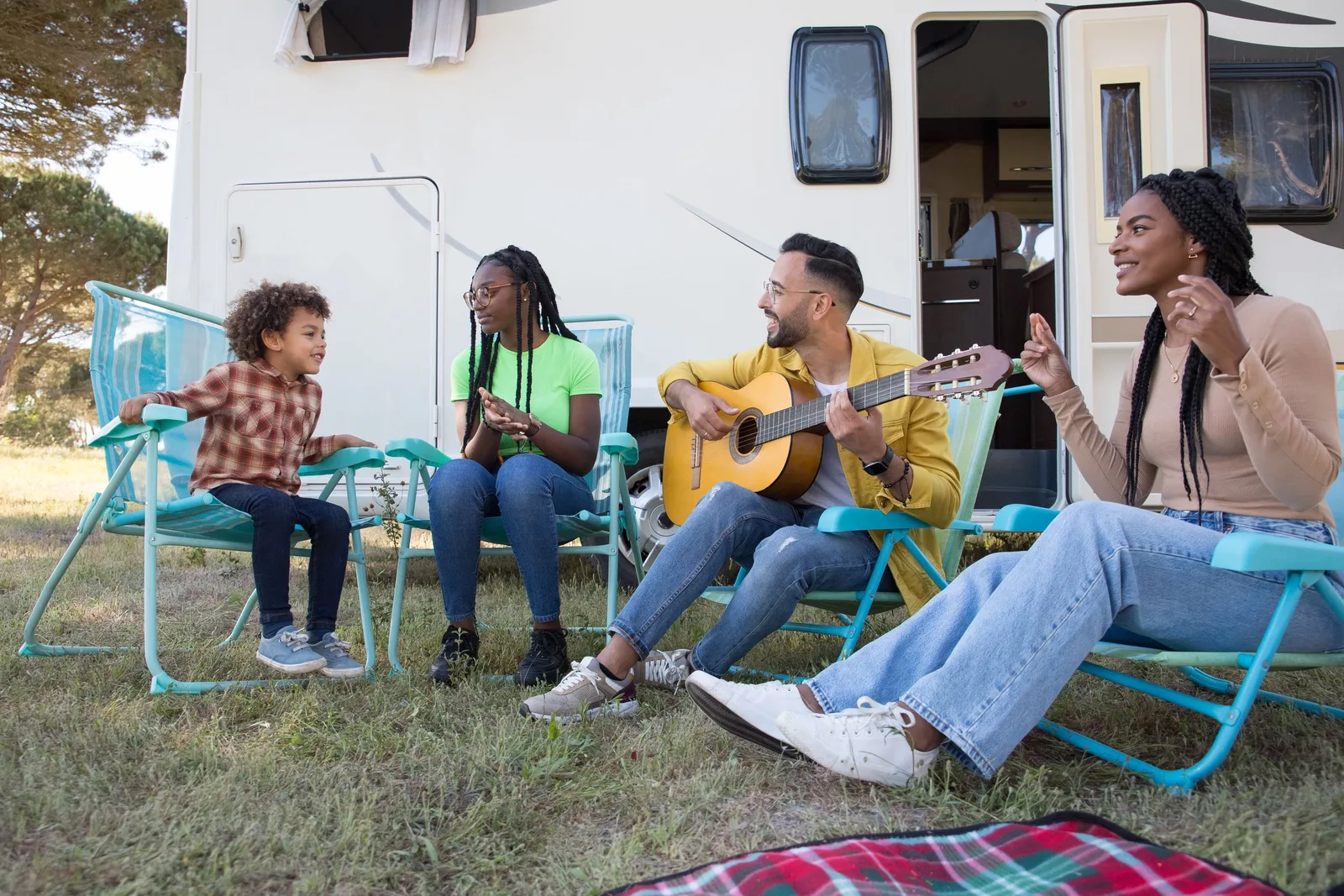 A family of four sits in teal folding chairs outside a camper van, with the father playing guitar and the mother singing.