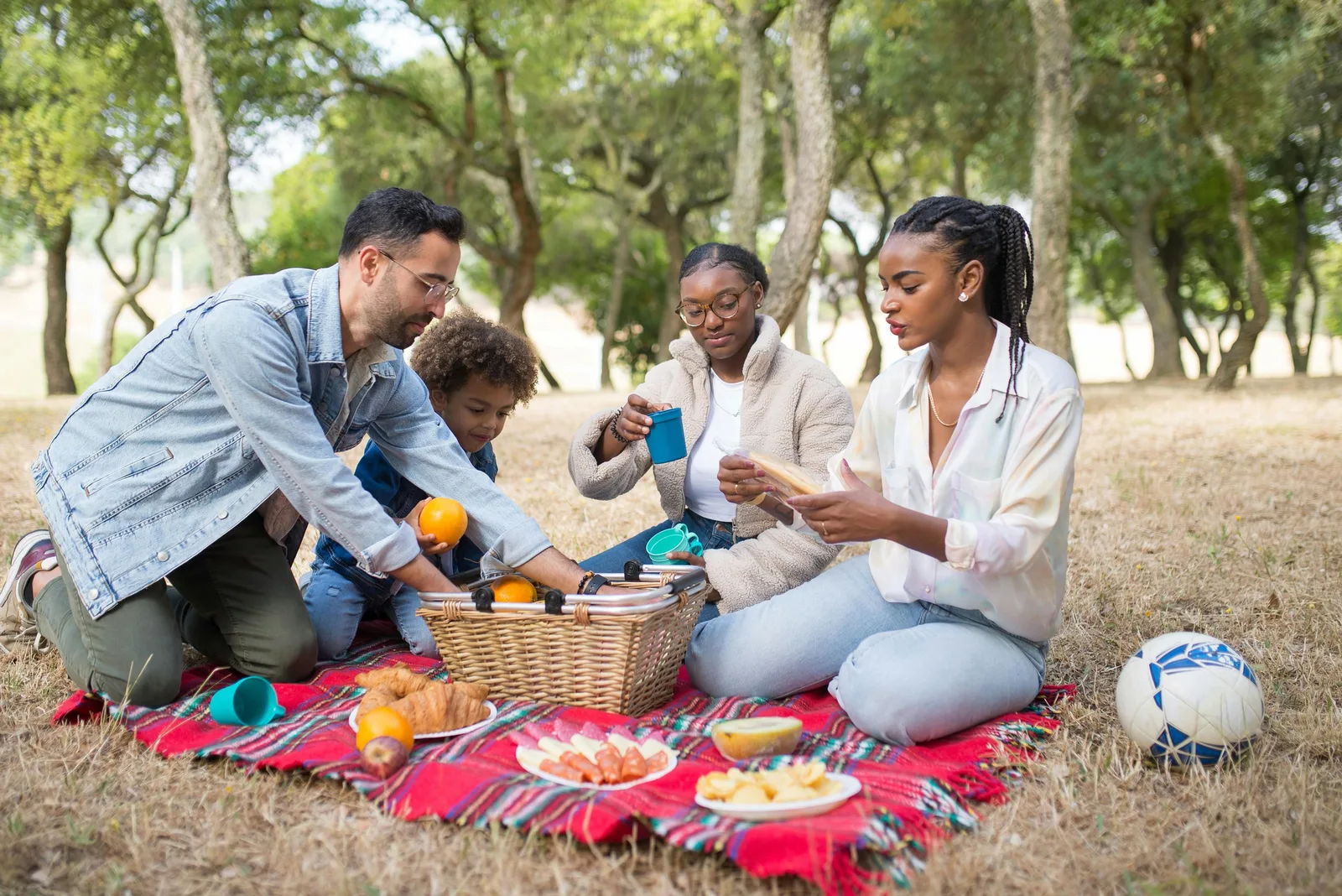 A family of four sits on a red picnic blanket in a park sharing food from a basket, with a soccer ball nearby.