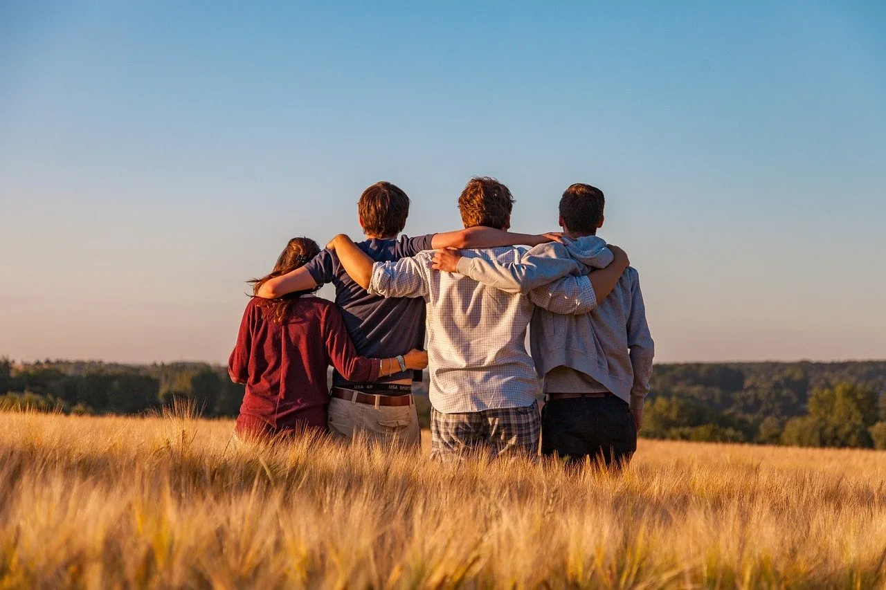 Four friends stand with their backs to the camera, arms around each other, looking out over a golden field at sunset.