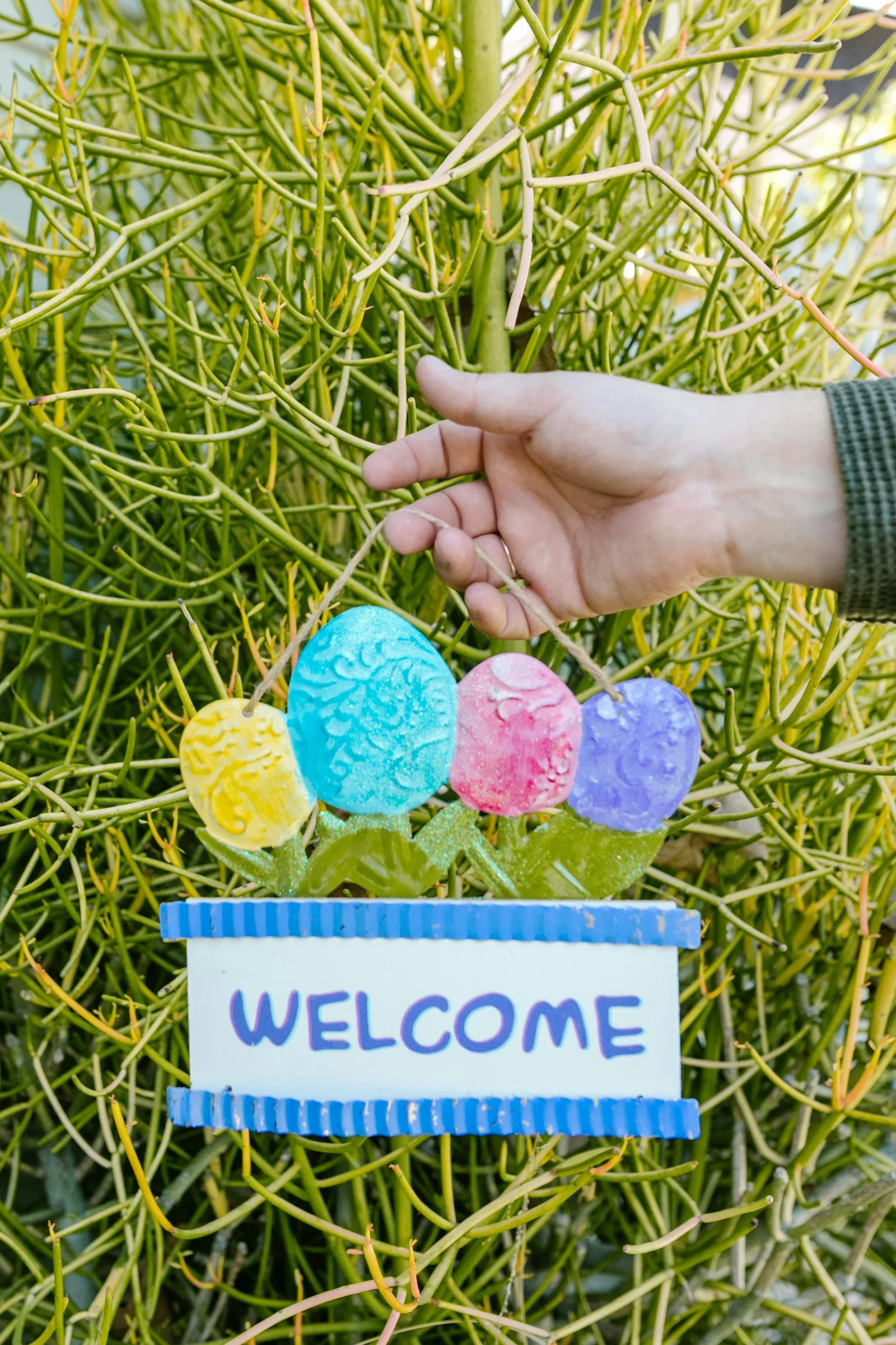 A hand places a handmade "Welcome" sign decorated with colorful eggs among green plants.