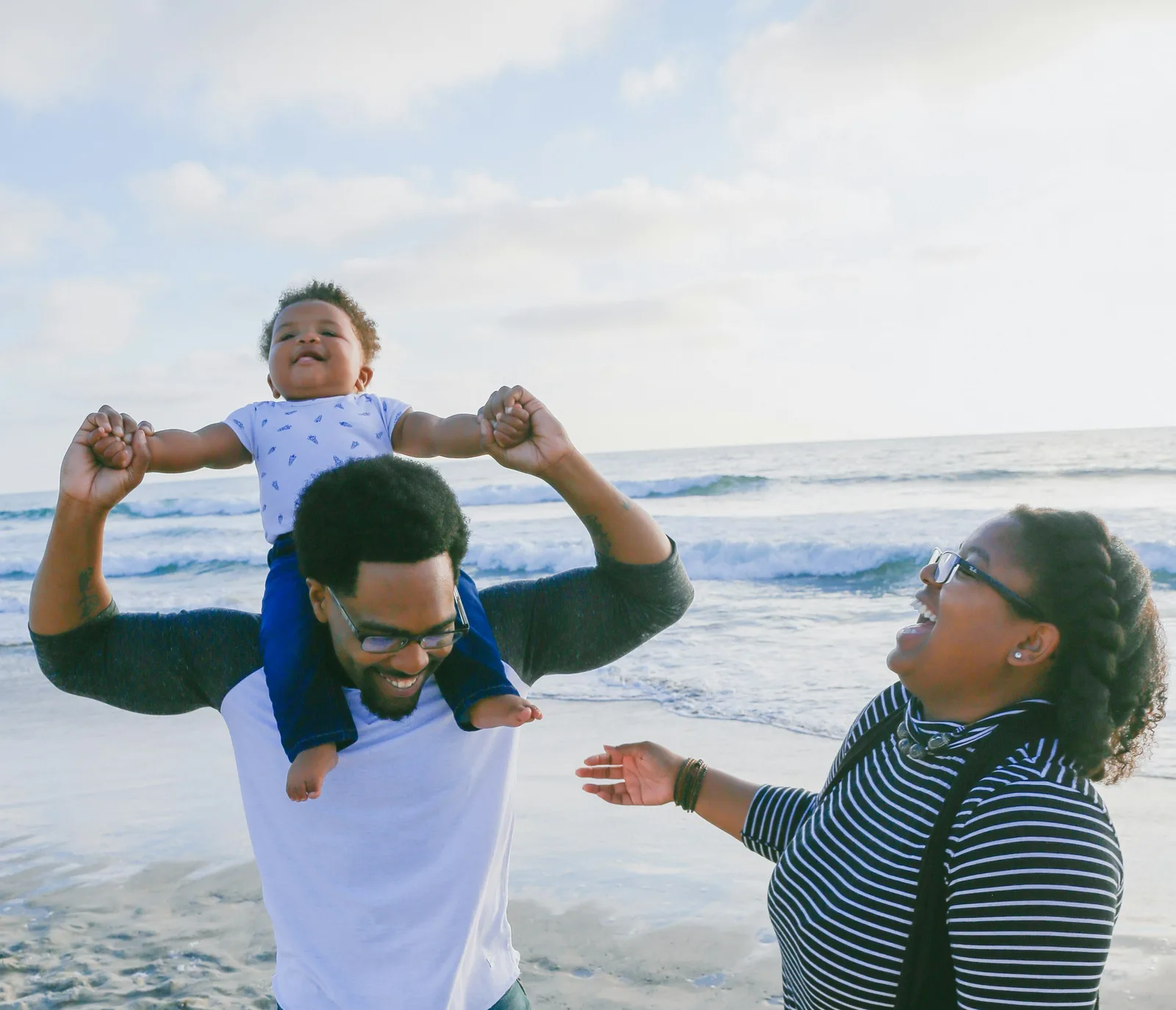 A father holds a laughing toddler up on his shoulders while a woman reaches toward them on a beach.