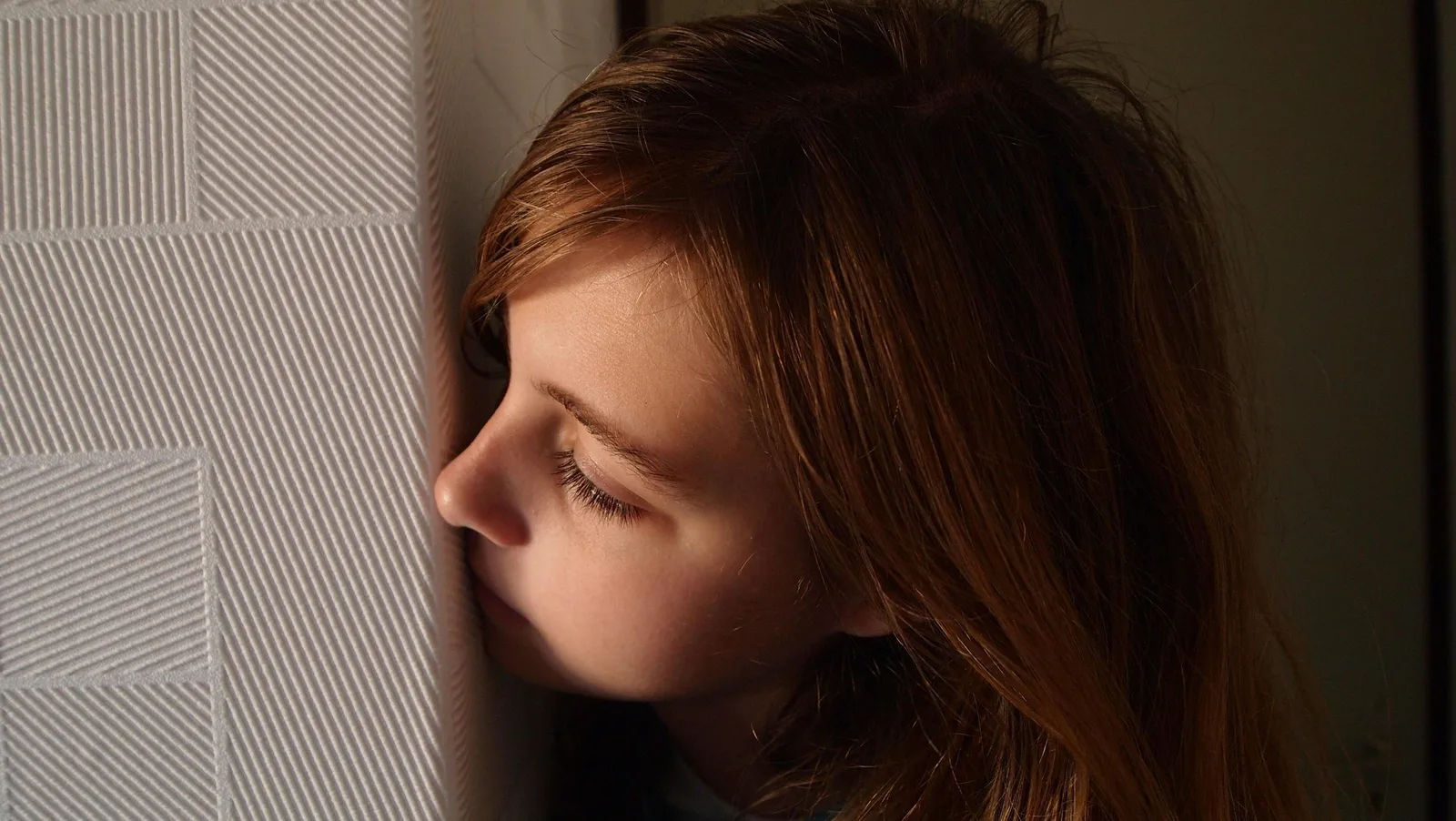 A young girl with long brown hair rests her forehead against a striped wall, looking down pensively.