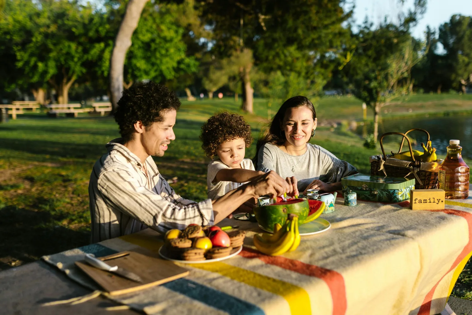 A family enjoys a picnic together at a park table covered with fruit, with a small sign reading "family" on the table.