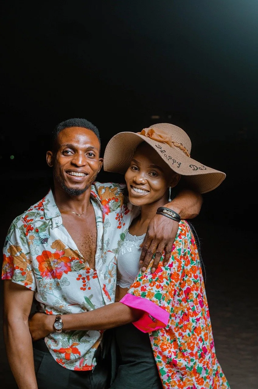 A couple smiles and embraces outdoors at night, the woman wearing a wide-brim sun hat.
