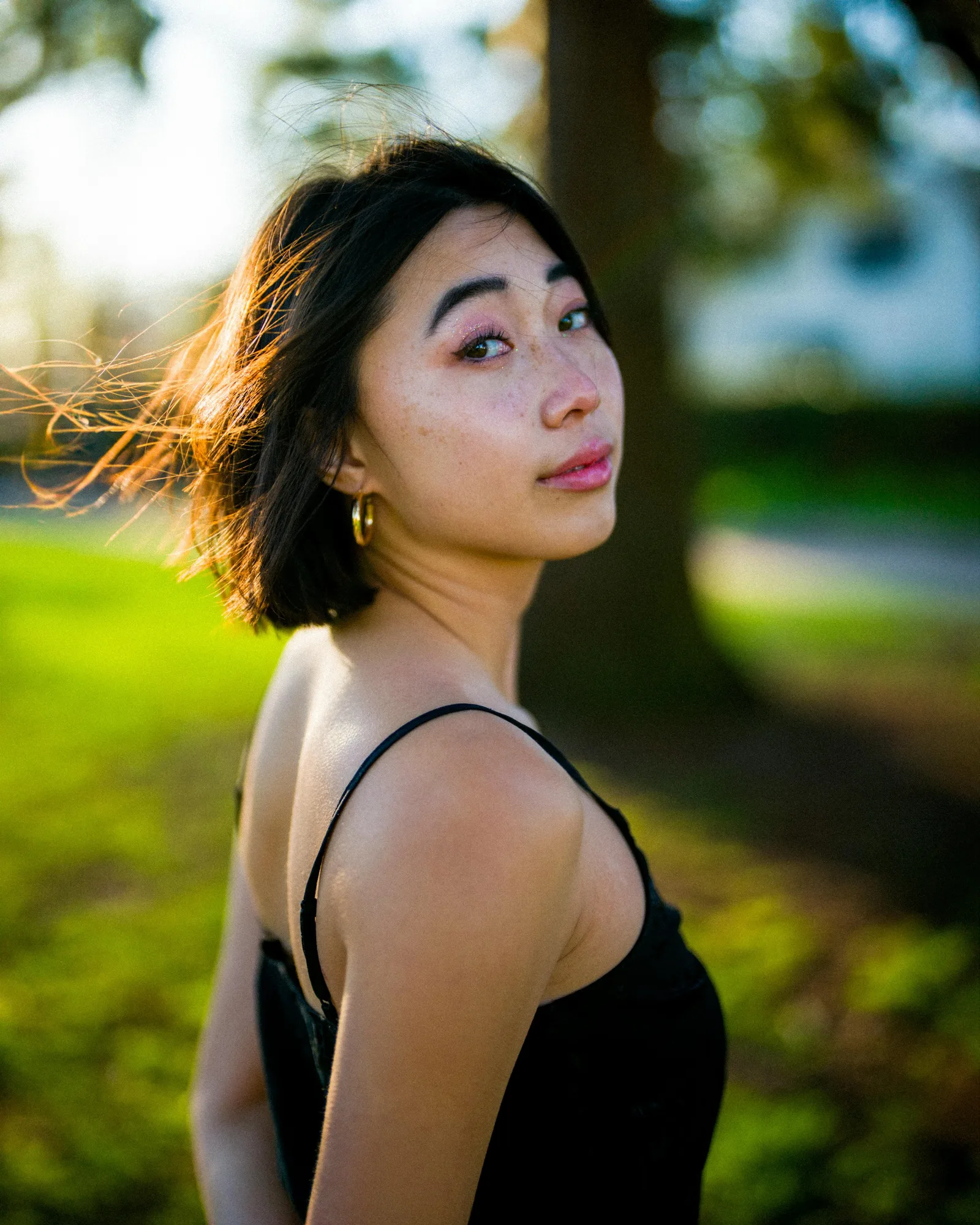 A young woman with short dark hair looks over her shoulder outdoors in golden-hour light.