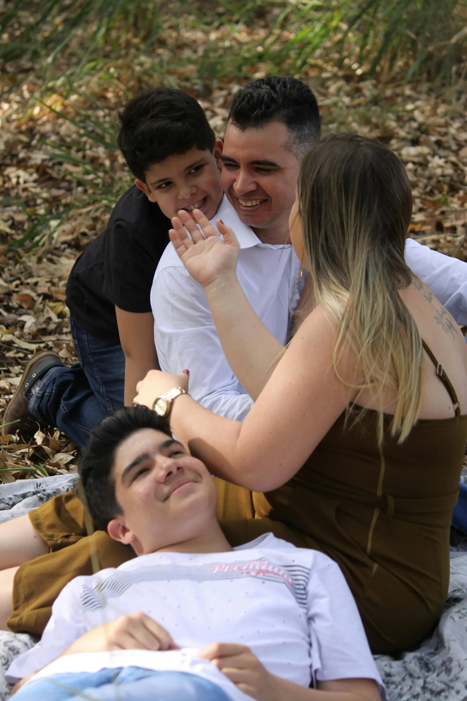 A family of four embraces and laughs together outdoors on a blanket among fallen leaves.