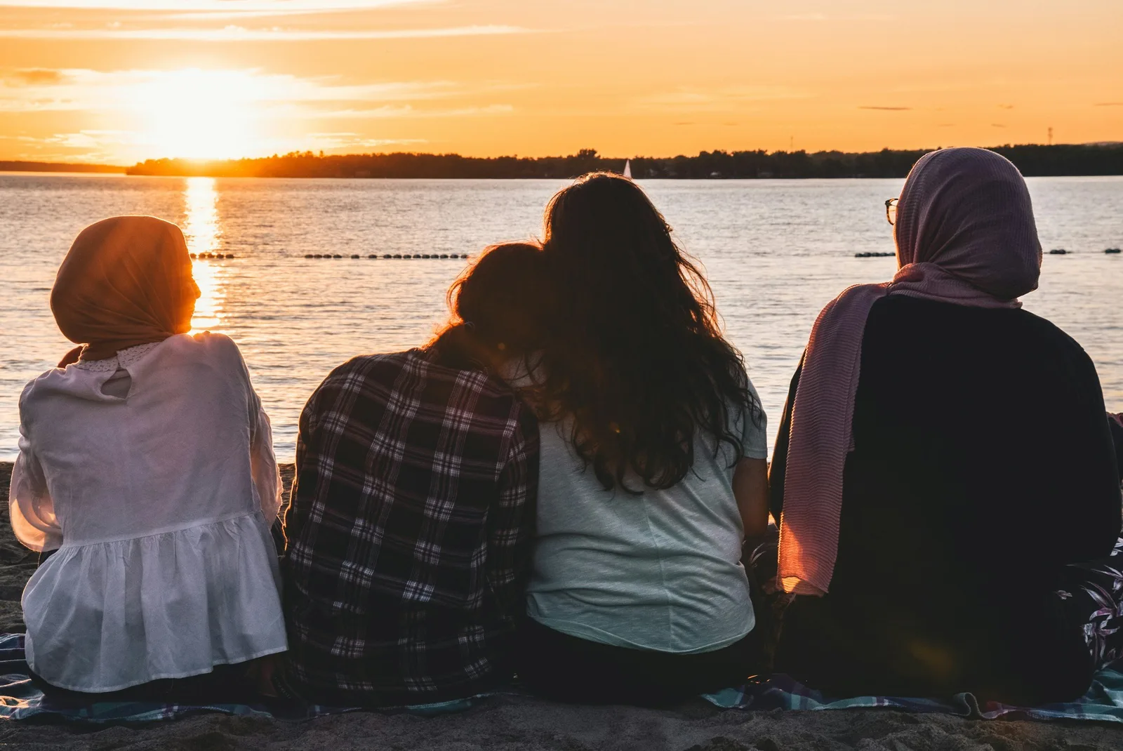 Four people in headscarves sit together watching the sunset over water.