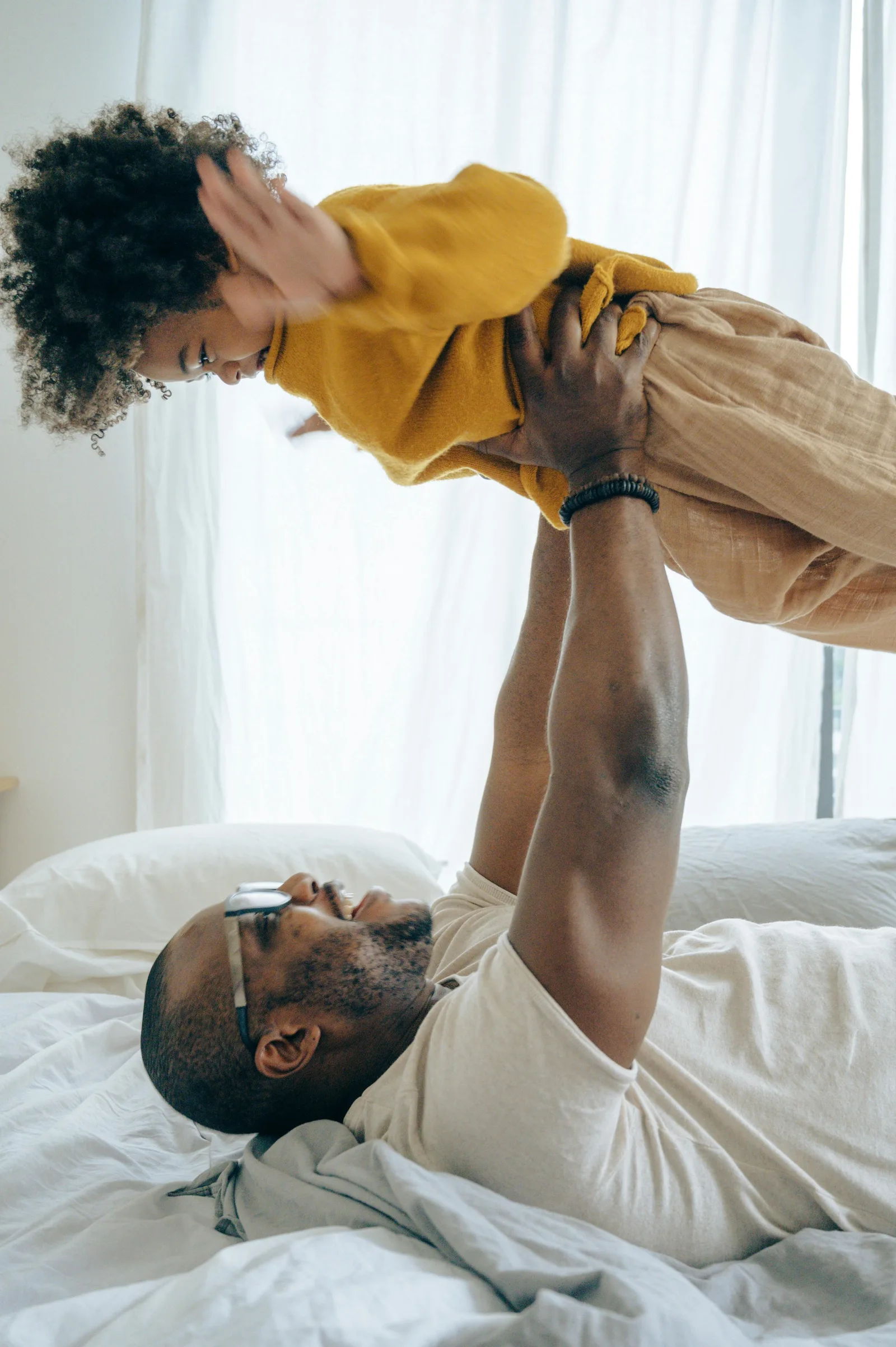 A father lying on a bed lifts a laughing toddler into the air above him.