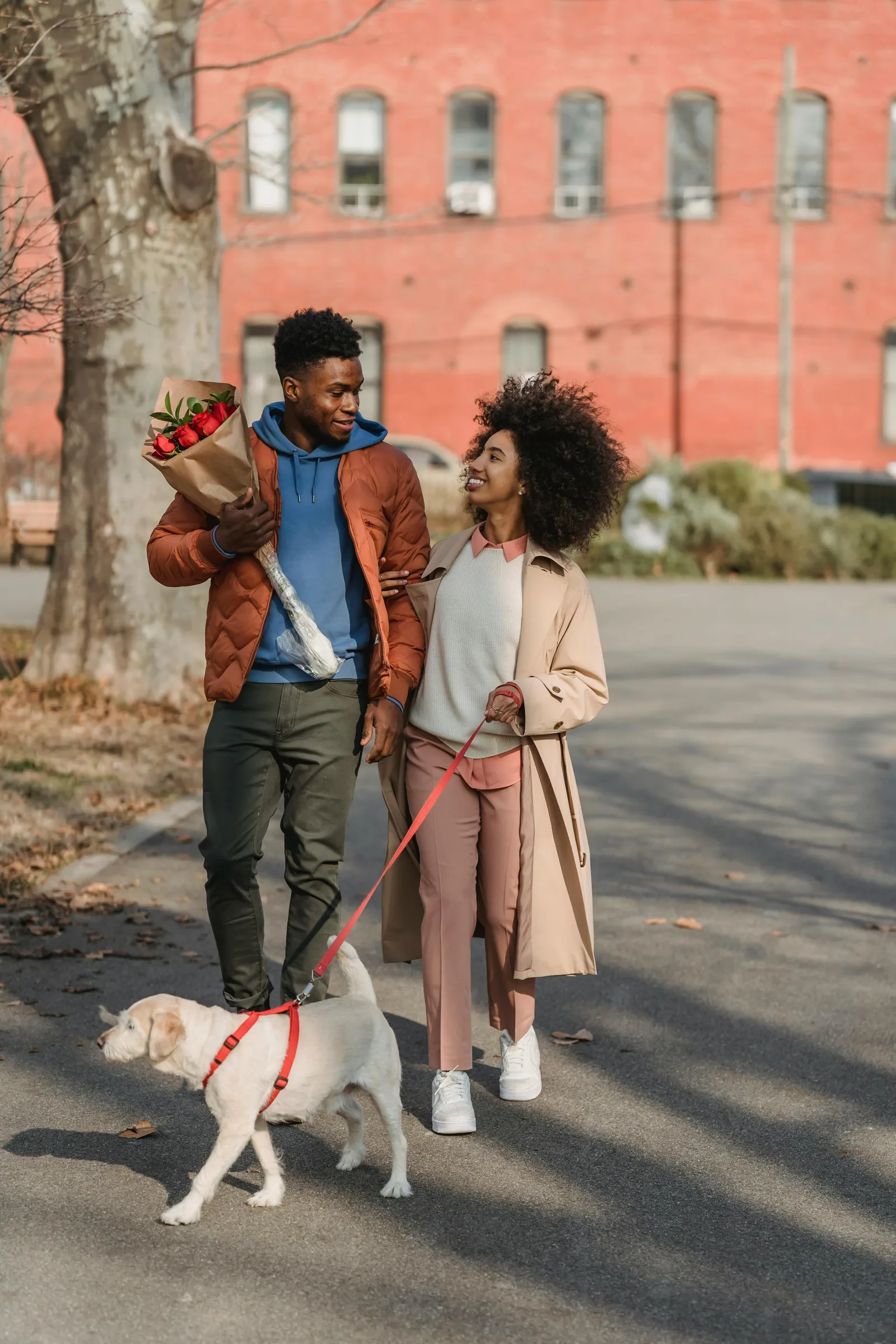 A couple walks a dog on a leash down a city sidewalk, the man carrying a bouquet of flowers.