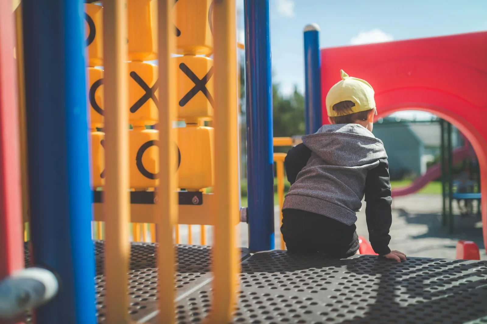 A young boy in a cap sits alone on a colorful playground structure, back to the camera.