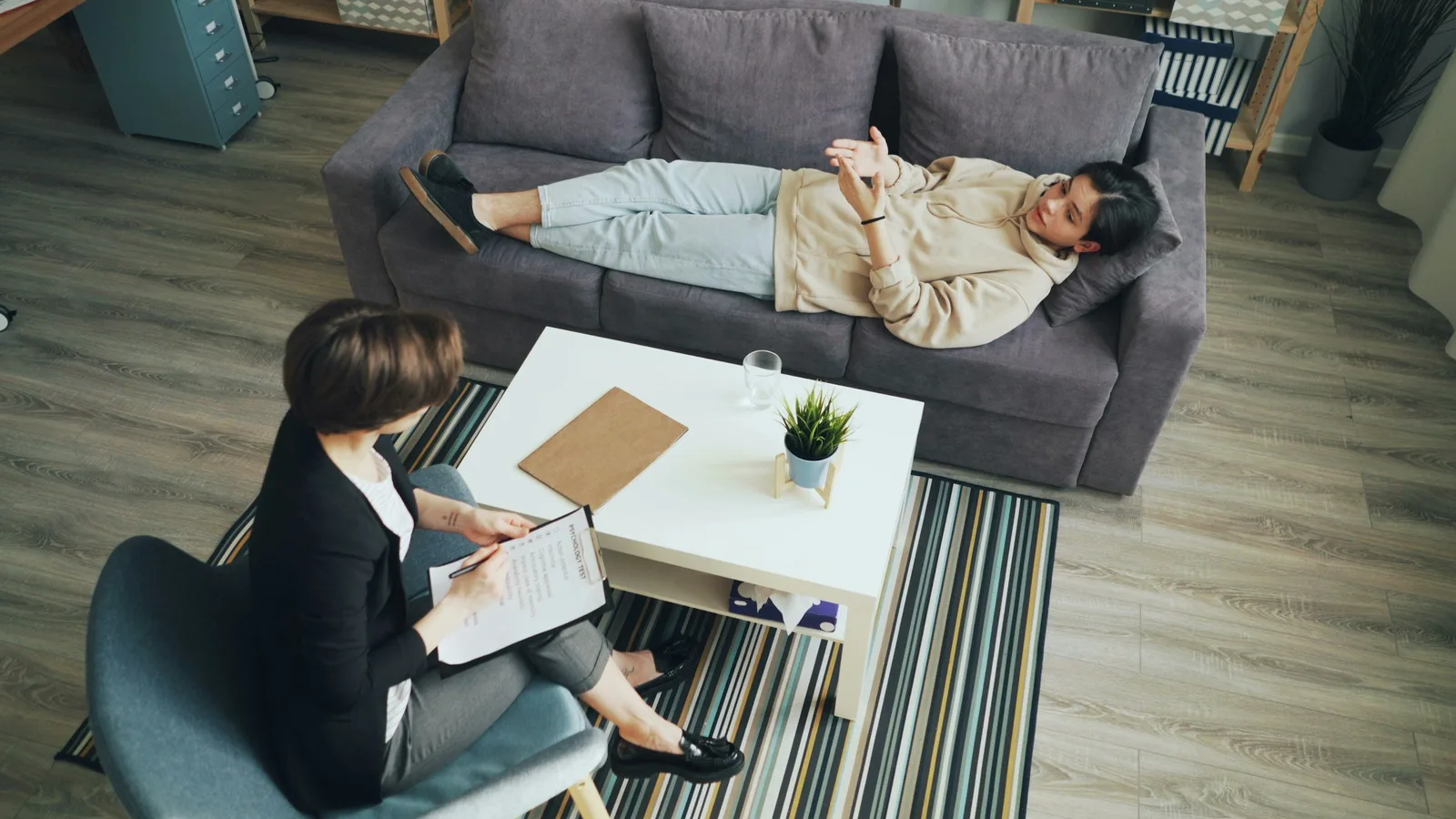 A therapist takes notes while a client reclines on a couch during a counseling session, viewed from above.
