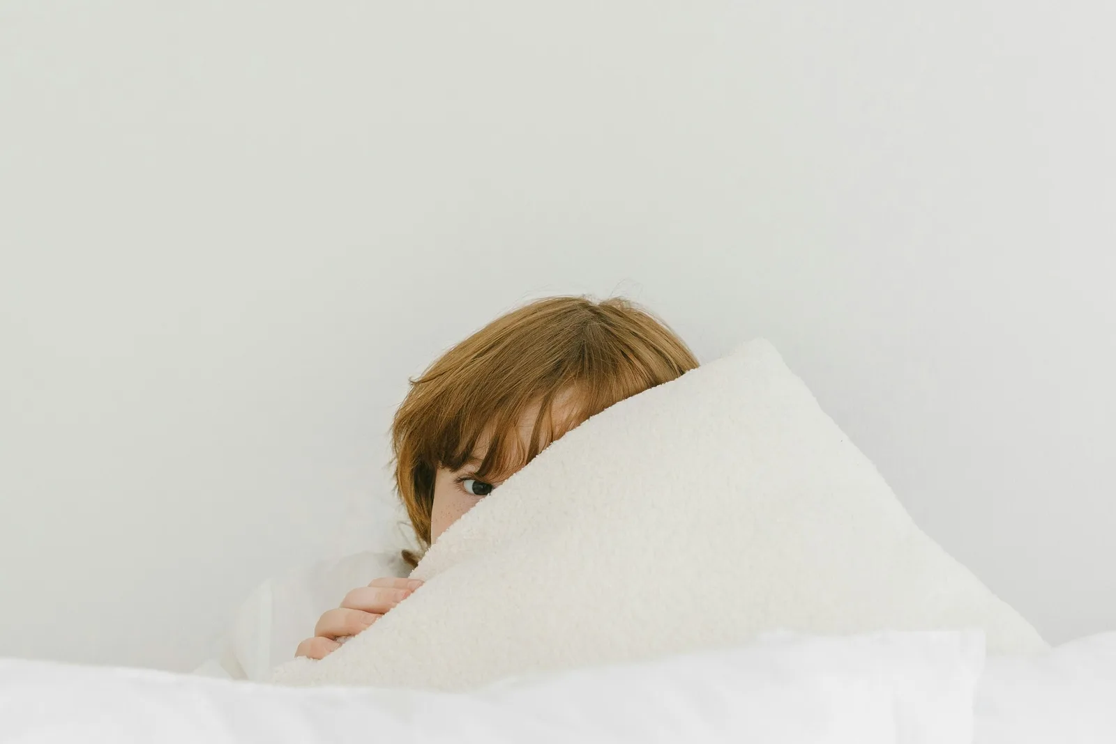 A person peeks over the top of a white blanket and pillow, only their eyes and hair visible.