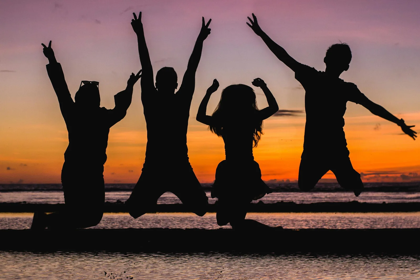 Silhouettes of four friends jumping and posing with raised arms against an orange sunset over water.