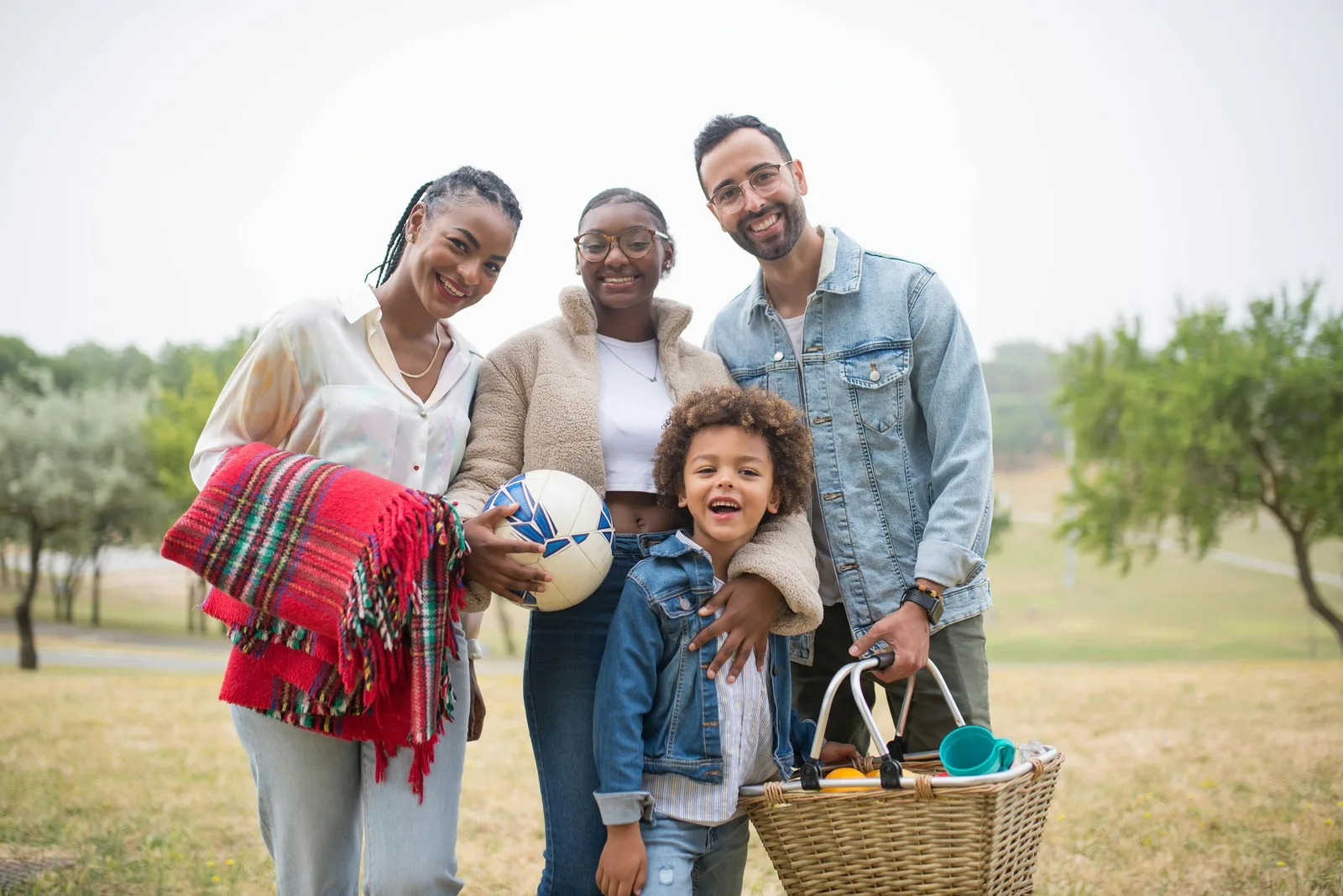 A family of four poses together outdoors holding a picnic blanket, a ball, and a basket.