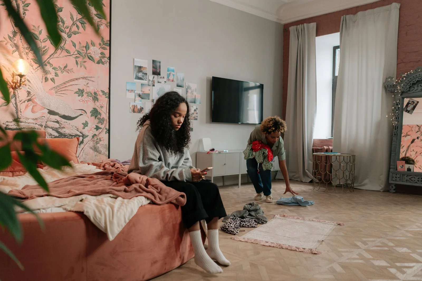 One woman sits on a bed looking at her phone while another tidies the bedroom floor.