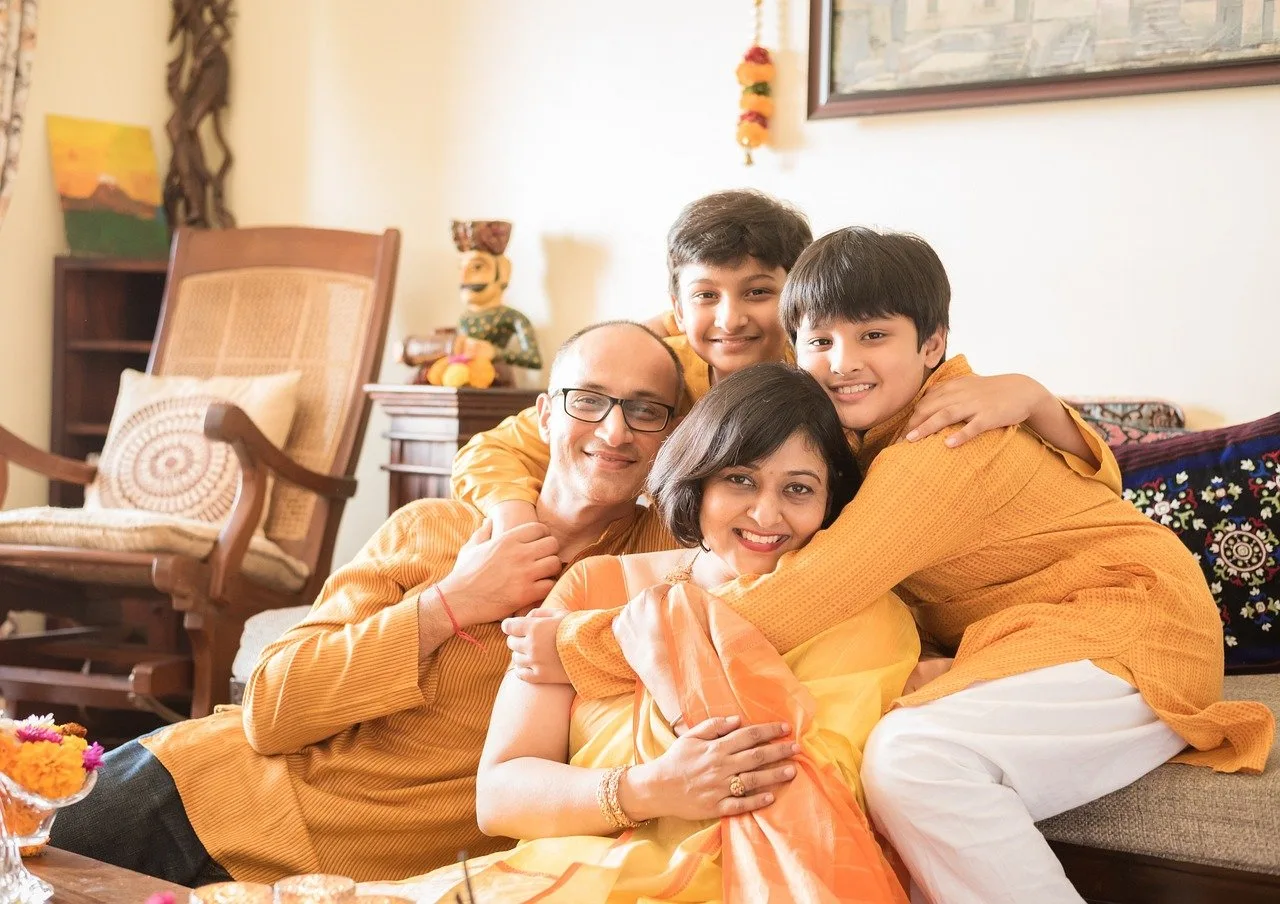 A family of four in matching orange and yellow traditional outfits embrace and smile together indoors.