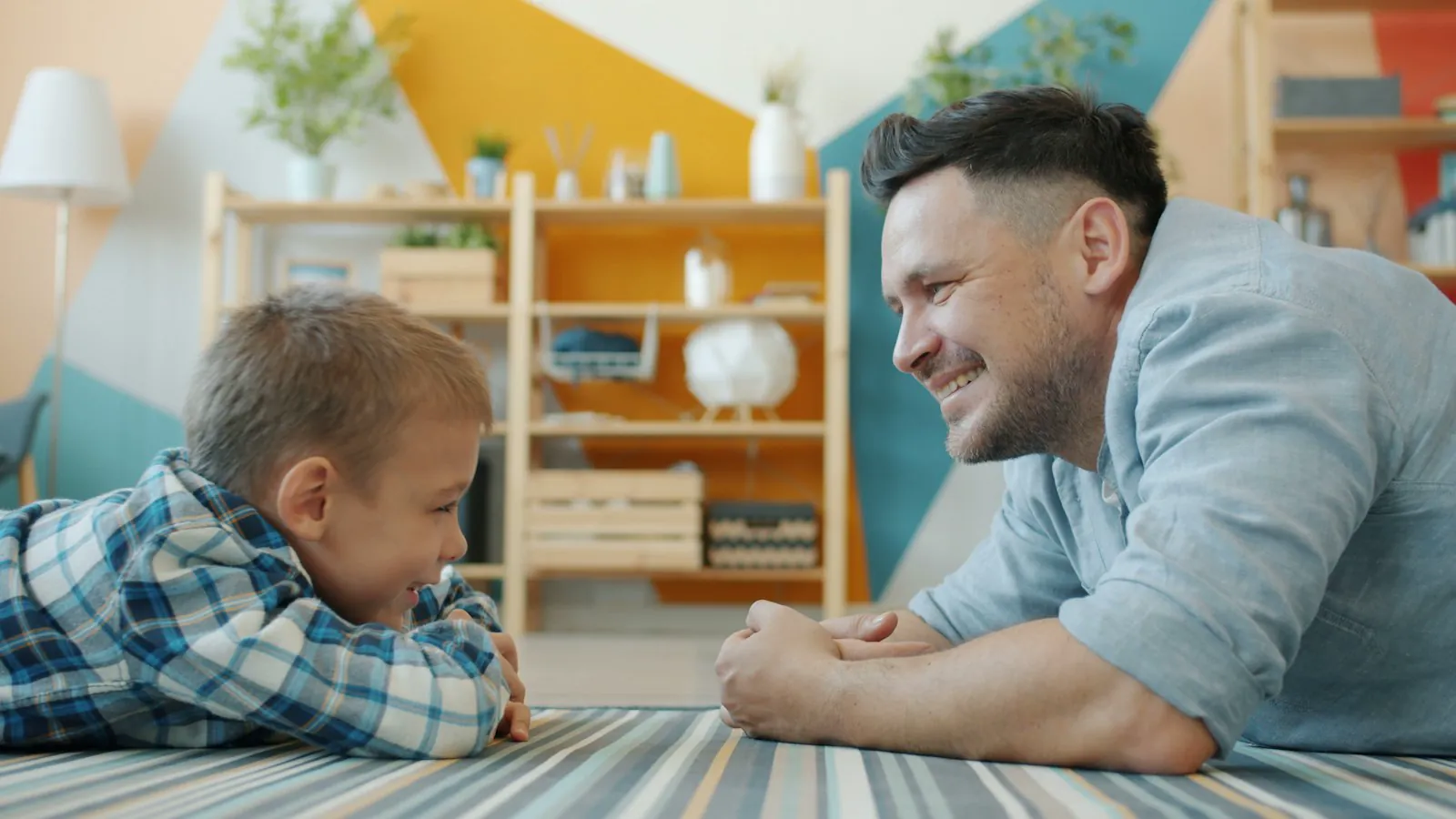 A father and his young son lie on the floor at home, talking together.