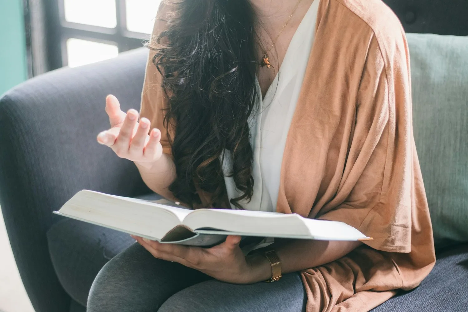 A woman with her face mostly hidden by her hair sits reading a book on a couch, gesturing with one hand.