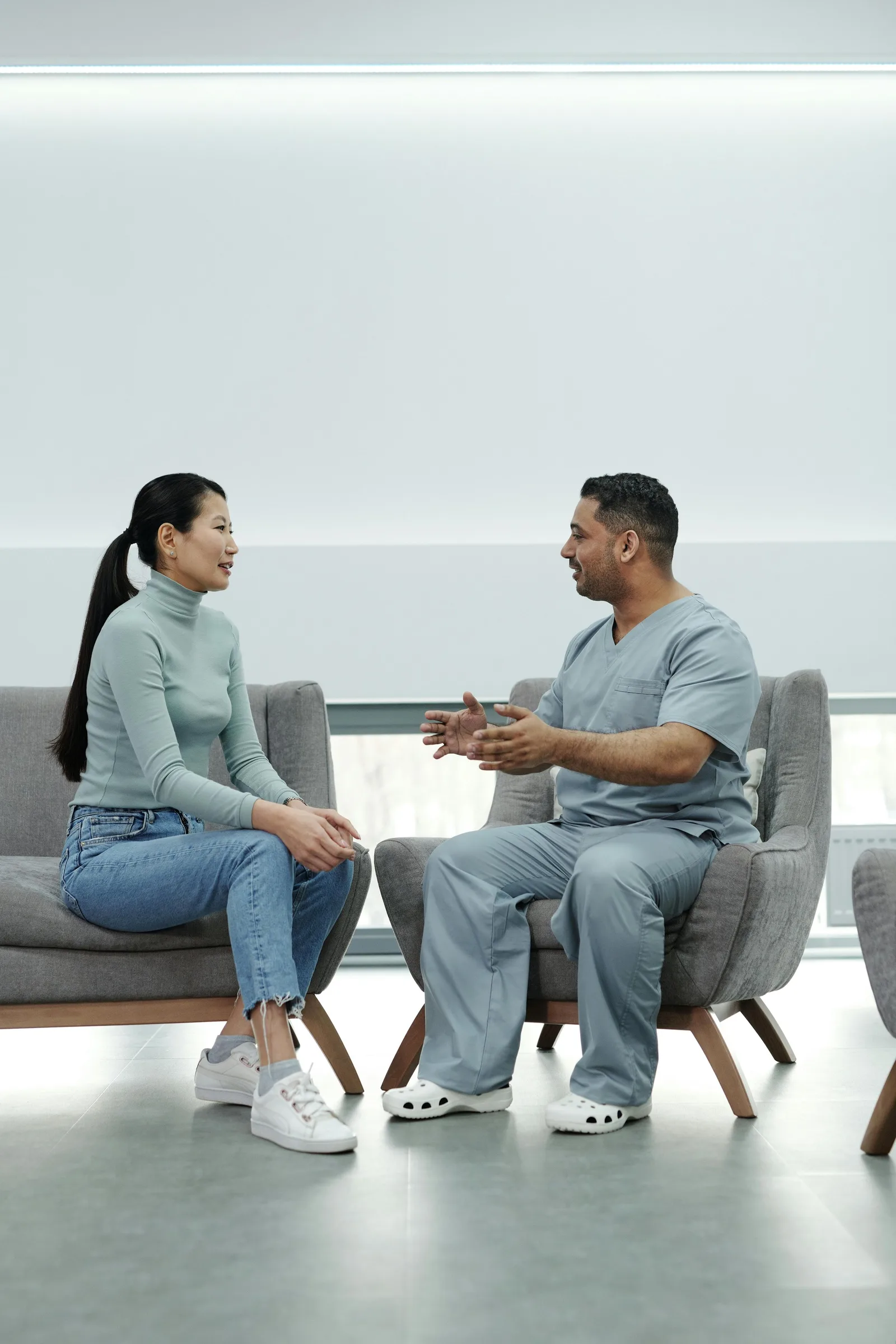 A clinician in scrubs talks with a seated woman in a waiting-room setting.