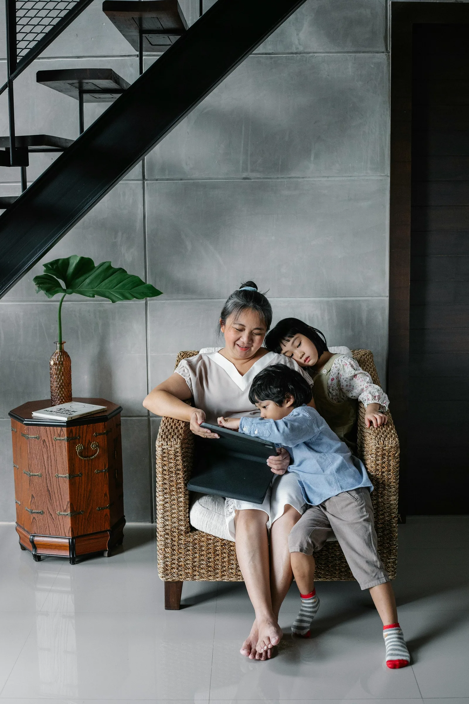 A grandmother sits in a chair with two grandchildren looking at a tablet together.