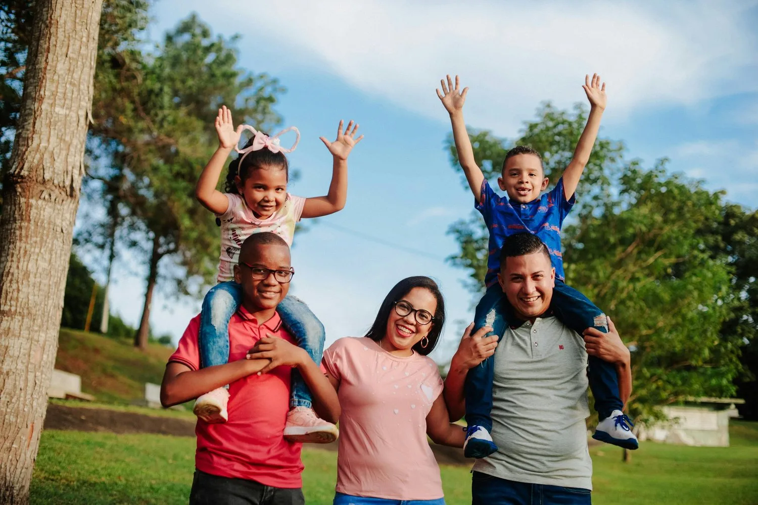 A family of five poses outdoors, with two children lifted onto the parents’ shoulders, all smiling with arms raised.
