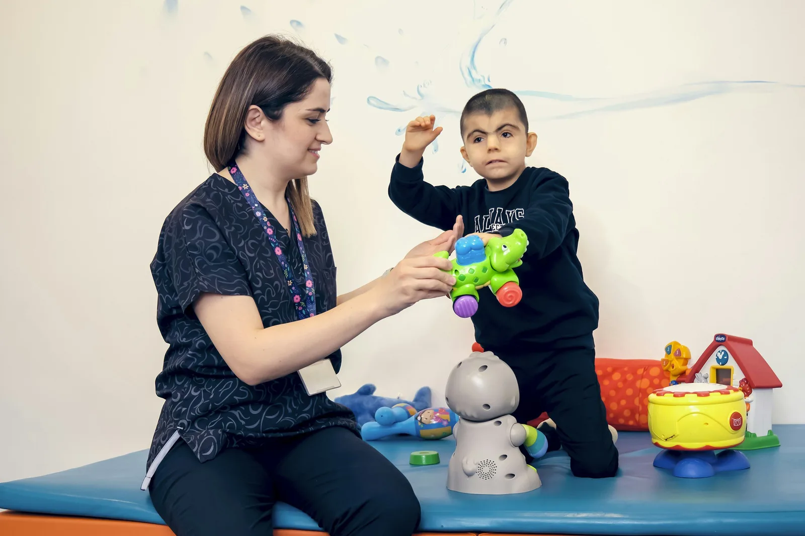 A clinician sits with a young child playing with toys on a therapy room mat.