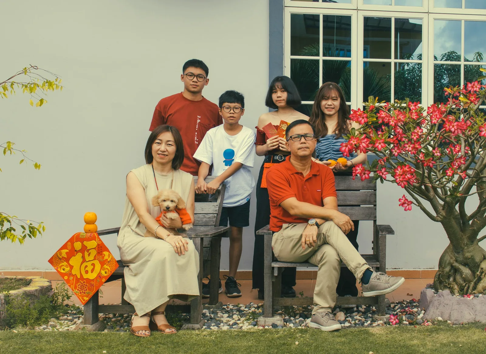 A family of six poses together outside a house decorated with a red lucky charm and a flowering tree, with a small dog.