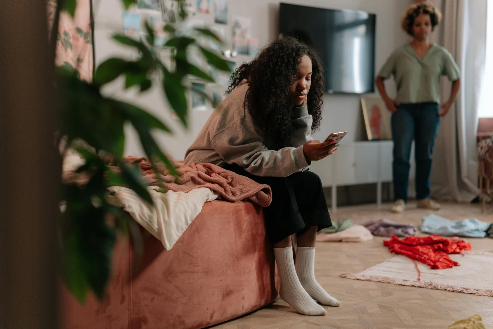 A girl sits on an ottoman looking at her phone while a woman stands in the background with hands on hips.