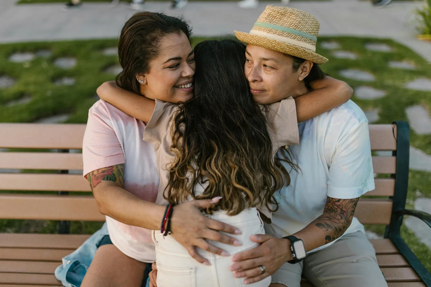 Three women embrace in a group hug on a park bench, one wearing a straw hat.