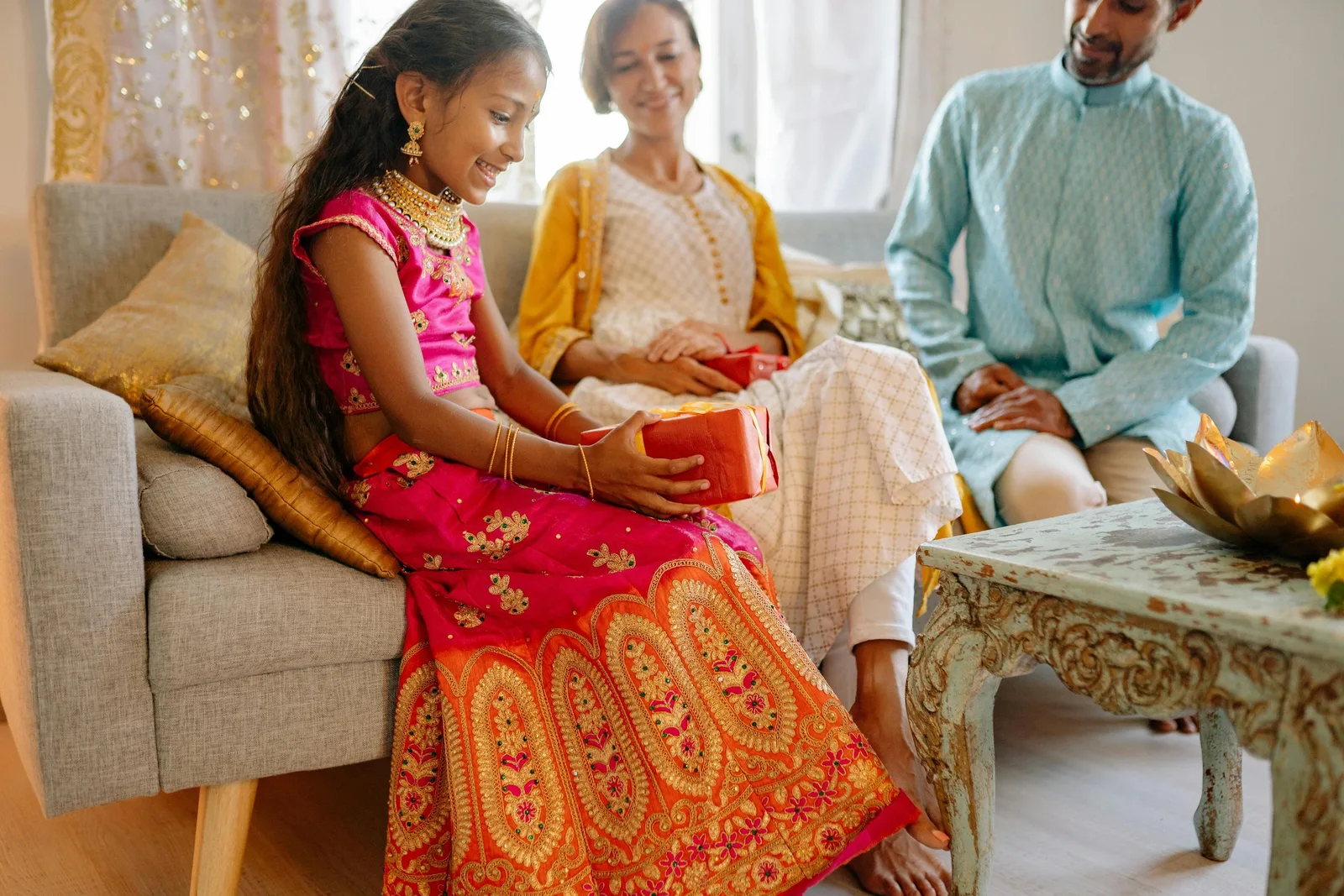 A girl in a bright pink and gold traditional outfit sits on a couch holding a gift box between two seated adults.
