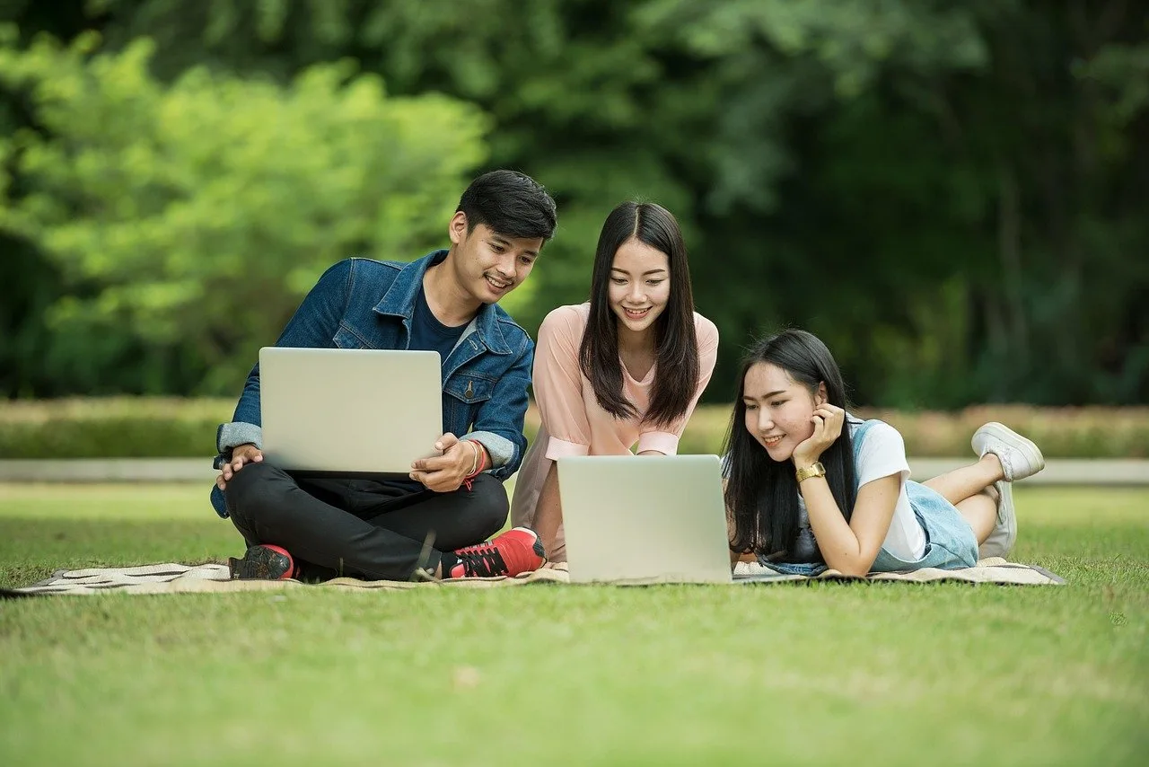 Three young people sit on a blanket in the grass looking at laptop screens together.