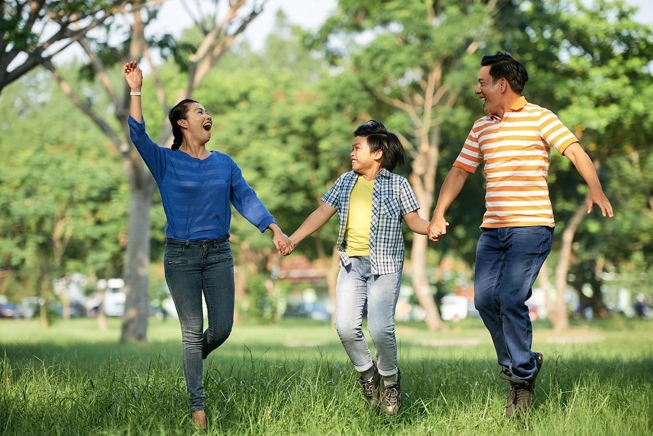 A mother, son, and father hold hands and laugh while walking through a park.