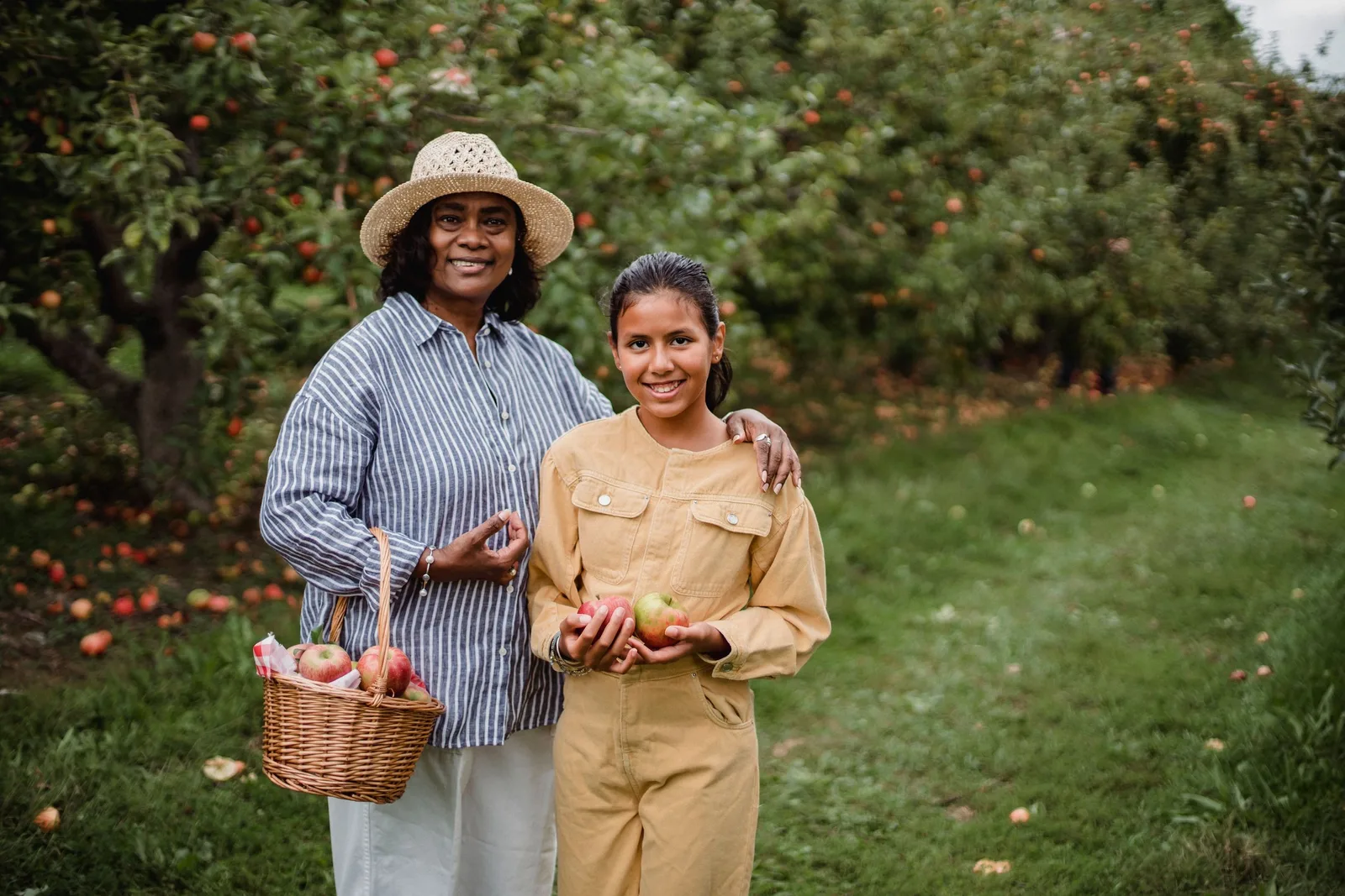 Two women stand together in an apple orchard, one holding a basket of apples, both smiling.
