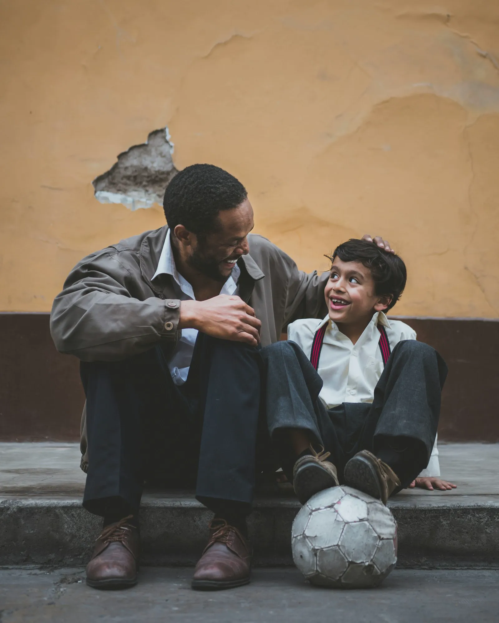 A father gently cradles his young son's head as the boy leans in holding a soccer ball.