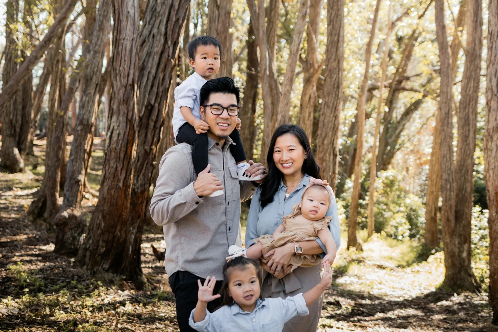 A family of five stands together in a forest, with a toddler on the father’s shoulders, a baby in the mother’s arms, and a young girl waving.