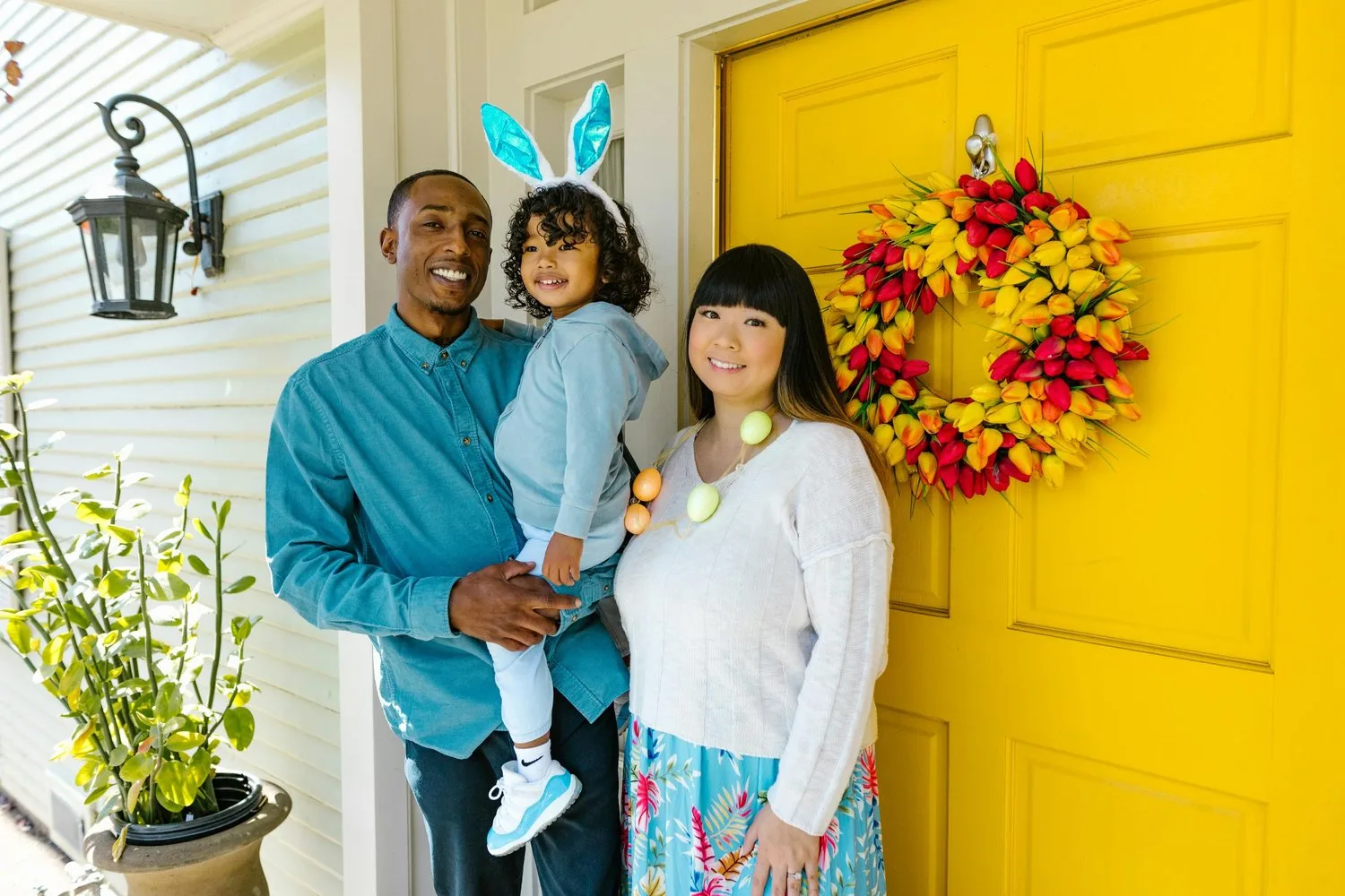 A man holds a toddler wearing bunny ears in front of a bright yellow door decorated with a floral wreath, standing beside a woman.