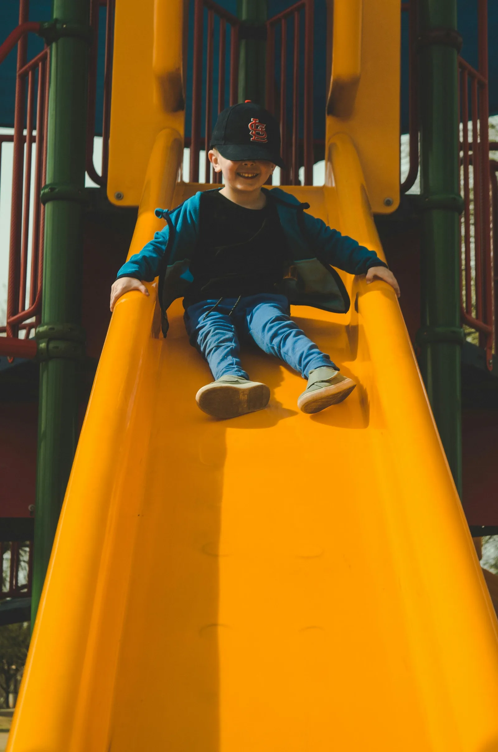 A boy in a cap smiles while sliding down a yellow playground slide.