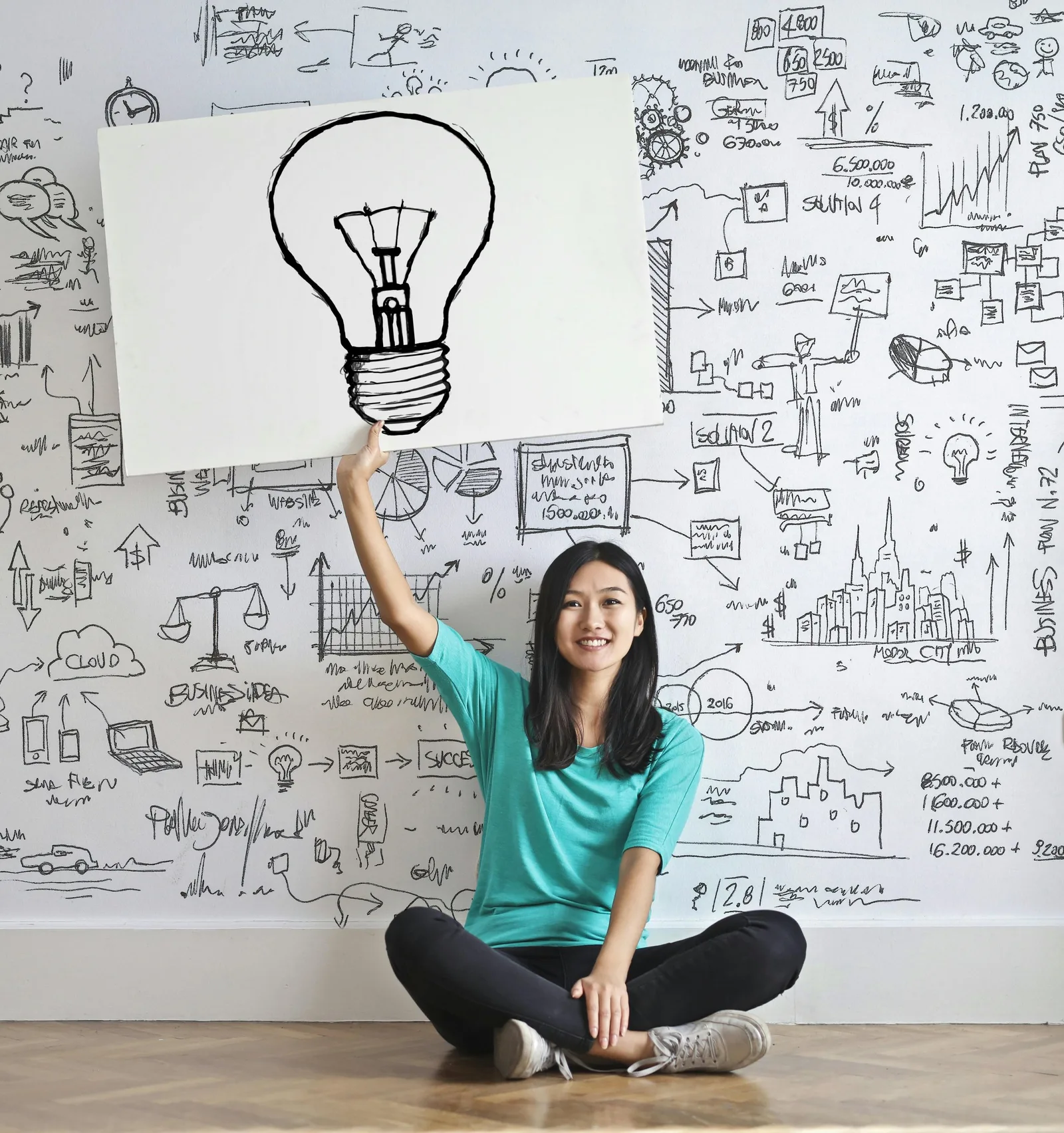 A woman sits cross-legged holding up a lightbulb drawing in front of a wall covered with idea sketches.