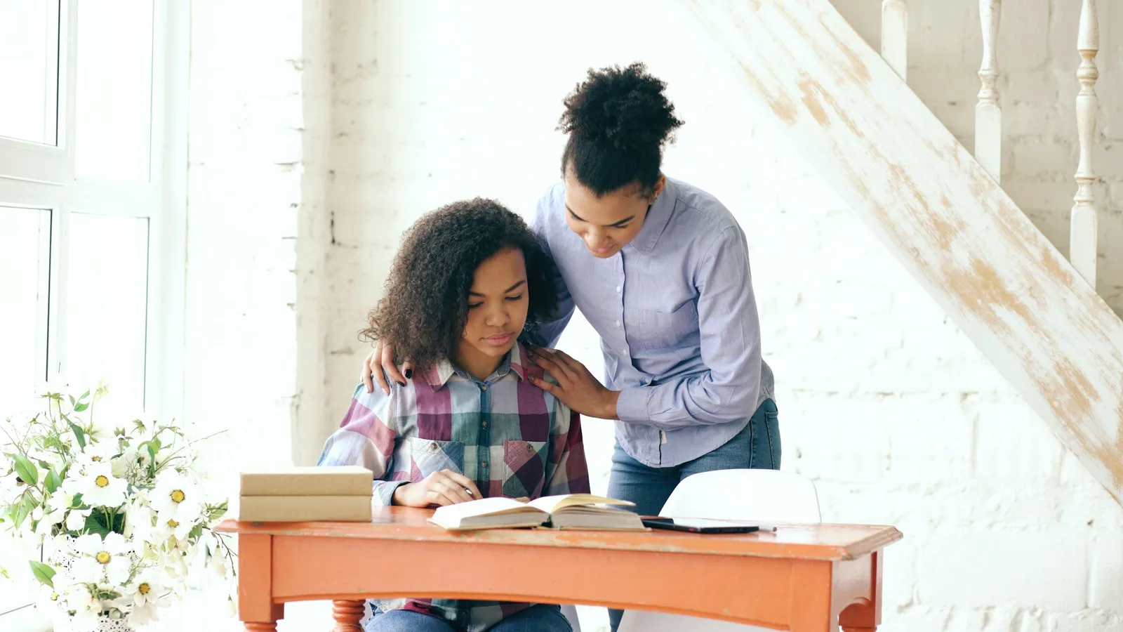 A teenage girl sits at a desk reading and writing while a woman stands behind her with a hand on her shoulder.