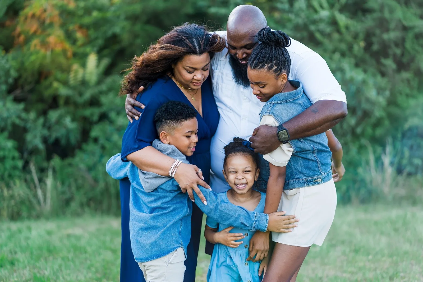 A family of five embraces closely outdoors on grass, smiling and laughing together.