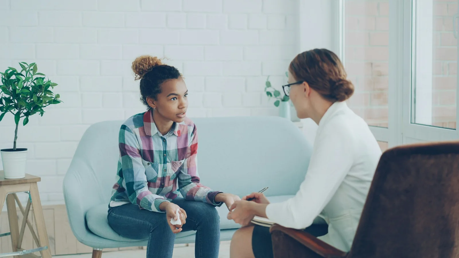 A teenage girl sits on a couch talking with a therapist who is taking notes.